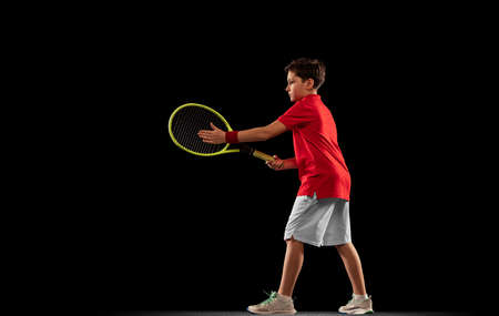 Full-length portrait of sportive young boy, playing tennis, training isolated over black backgound.の写真素材