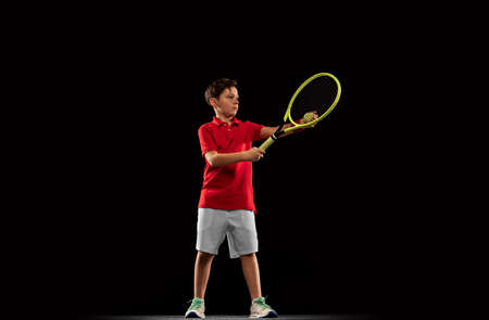 Full-length portrait of young boy, tennis player in motion, training, serving ball isolated over black backgroundの写真素材