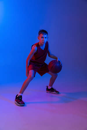 Full-length portrait of boy, basketball player in motion, training isolated over gradient blue purple background in neon lightsの写真素材
