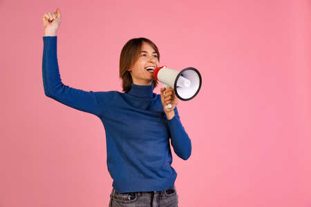 Cropped portrait of excited young woman shoouting in megaphone isolated over pink background. News spreadingの写真素材