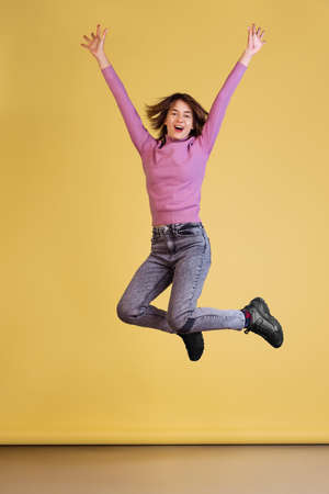 Full-length portrait of young woman with cheerful emotions wearing casual clothes jumping isolated over yellow backgroundの写真素材