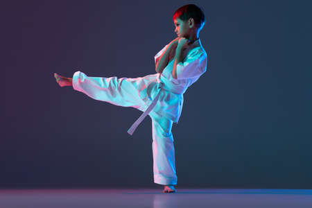 Full-length portrait of sportive child, training karate punching isolated over gradient blue background in neonの写真素材