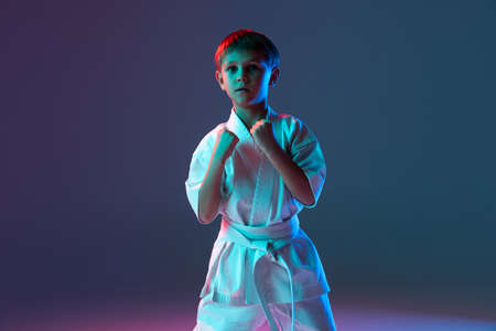 Portrait of little boy in white kimono training karate isolated over gradient blue background in neon. Fists upの写真素材