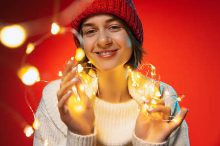Cropped portrait of young happy woman holding holiday lights, decorations isolated over red backgroundの写真素材