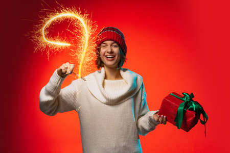 Cropped portrait of young joyful woman holding present and holiday sparkles isolated over red...