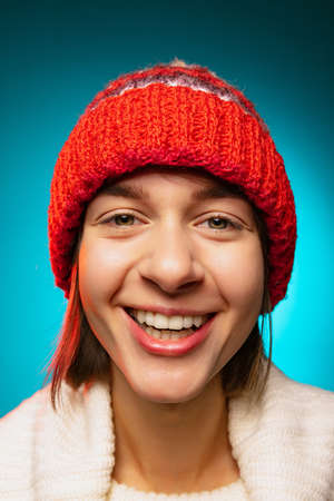 Cropped portrait of young merry girl in red winter hat smiling at camera isolated over blue backgroundの写真素材