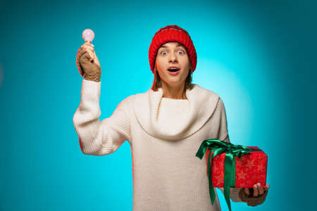 Cropped portrait of young girl in red winter hat with big present box and lightbulb posing isolated blue background. Ideasの写真素材