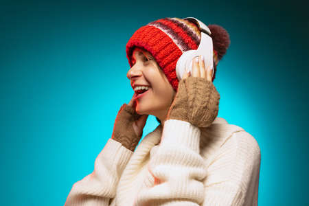 Cropped portrait of young joyful girl listening to cheerful music in white headphones isolated over blue background. Side viewの写真素材