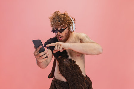 Cropped portrait of man in character of neanderthal using phone and listening to music in headphones isolated over pink backgroundの写真素材