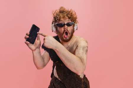 Portrait of man in character of neanderthal in headphones pointing at phone with shocked expression isolated over pink backgroundの写真素材