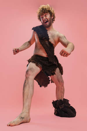 Full-length portrait of man in character of neanderthal wearing animal skin cloth and showing crazy...