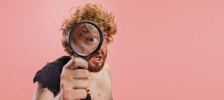 Cropped portrait of man in character of primitive person, neanderthal holding magnifier enlarging eye isolated over pink backgroundの写真素材