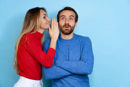 Cropped portrait of happy couple, woman whispering to shocked man gossips, news isolated over blue backgroundの写真素材