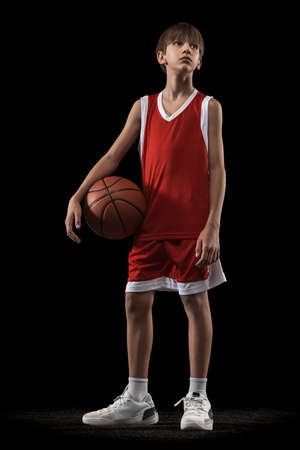 Full-length portrait of teen boy, basketball player in red uniform posing with ball isolated over black backgroundの写真素材