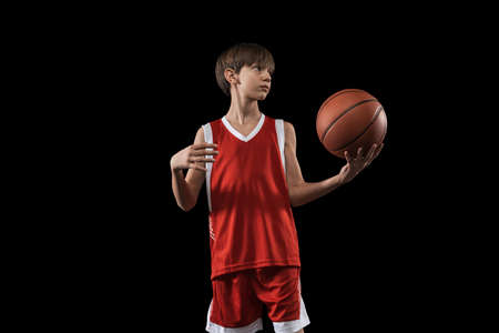 Cropped portrait of young boy, basketball player in red uniform standing, posing with ball isolated over black backgroundの写真素材