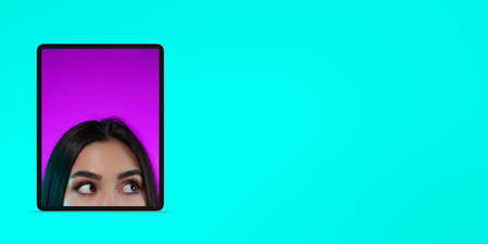 Creative design. Art collage. Half-face portrait of young beautiful girl, eyes looking out phone screen isolated over blue backgroundの写真素材