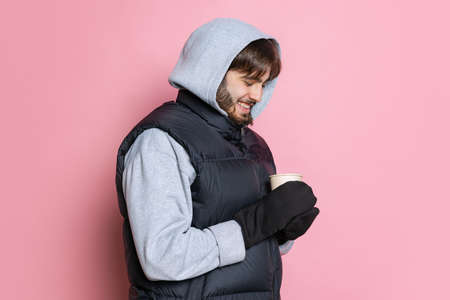 Cropped side view portrait of young man waring hood and vest, holding ea cup isolated over pink...
