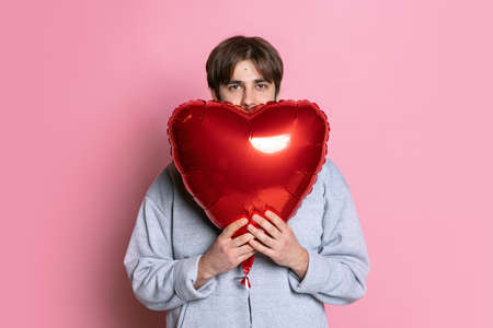 Cropped portrait of young boy covering face with red balloon in shape of heart isolated over pink studio backgroundの写真素材