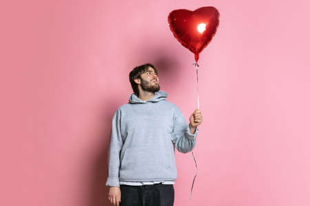 Portrait of young boy holding red balloon in heart shape isolated over pink background. Valentines Dayの写真素材