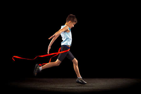 Full-length portrait of boy, child crossing red ribbon isolated over black background. Winning...