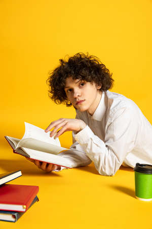 Portrait of young boy, student lying on floor, reading book, doing homework isolated over yellow...