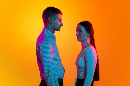 Portrait of two young people, man and woman in white cloth calmly standing, posing isolated over orange background in blue neonの写真素材