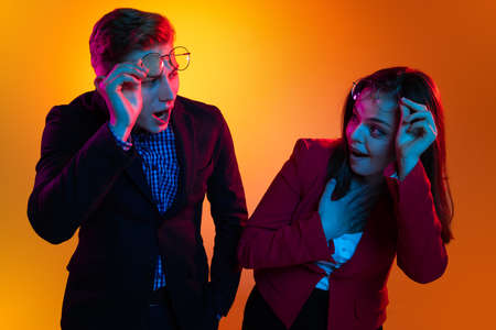 Portrait of young man and woman rising glasses and looking at each other with shock isolated over...