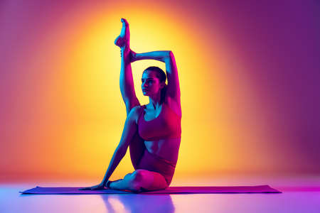 Portrait of young sportive girl doing yoga exercises, sitting, rising leg upwards isolated over...