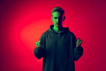 Half-length portrait of young man in black hoodie with serious expression isolated over red background in green neonの写真素材