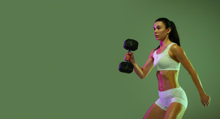 Portrait of young muscular sportive girl training, lifting dumbbell isolated over green studio background in neon light. Concept of sport, fitness, healthy lifestyle, strength, youth, adの写真素材