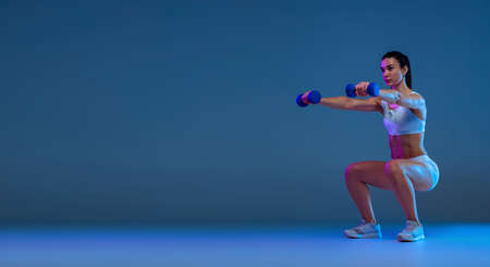 Portrait of young sportive girl doing squats with dumbbell, training isolated over blue studio...