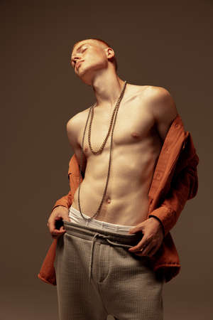 Portrait of young stylish red-haired man posing in unbuttoned oramge shirt, wearing trendy necklace isolated on grey studio background. Concept of fashion, style, body aesthetics, beauty, mens healthの写真素材