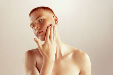 Portrait of young red-haired man with freckles touching cheeks, posing isolated over grey studio background. After shaving. Concept of mens health, lifestyle, beauty, body and skin careの写真素材