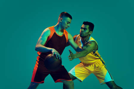 Portrait of two young men, basketball players in uniform training isolated over blue background in neon light. Defender position. Concept of sport, team game, action, active lifestyle, adの写真素材