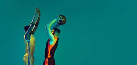 Portrait of two young men, professional basketball players in a jump, throwing ball into basket isolated over blue background in neon light. Concept of sport, team game, action, active lifestyle, adの写真素材