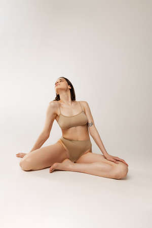 One beautiful young woman sitting on floor, posing in cotton beige underwear isolated on grey studio background. Sensuality. Concept of beauty, body and skin care, wellness, health, spa, cosmetics, adの写真素材