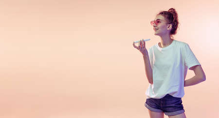 Portrait of young girl in white T-shirt recording voice message on phone, posing isolated over pink studio background in neon light. Concept of youth, beauty, lifestyle, fashion, fun, emotionsの写真素材