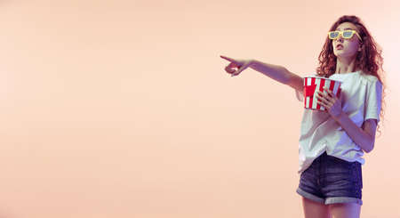 Portrait of young girl in casual outfit posing with popcorn basket isolated over pink studio background in neon light. SHowing emotions. Concept of youth, beauty, lifestyle, fashion, fun, emotionsの写真素材