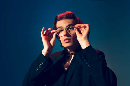 Portrait of stylish young man wearing official outfit with glasses, posing isolated over dark blue...