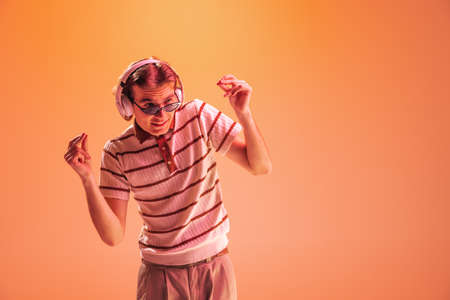 Portrait of young man listening to music in headphones, dancing isolated on orange studio background...
