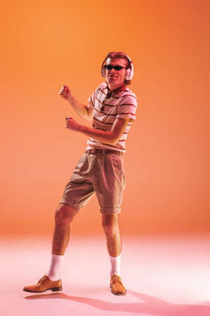 Portrait of young emotive man listening to music in headphones, dancing isolated over orange studio...