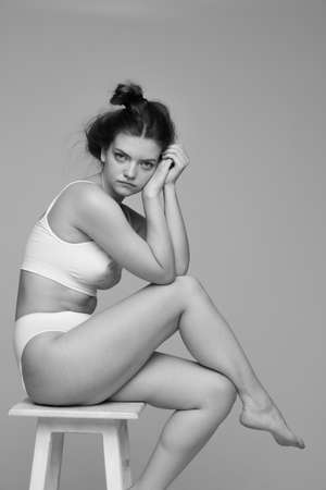One tender young girl with hair bun sitting on chair in white underwear, posing. Black and white photography. Concept of natural beauty, body and skin care, femininity, self-acceptance, adの写真素材
