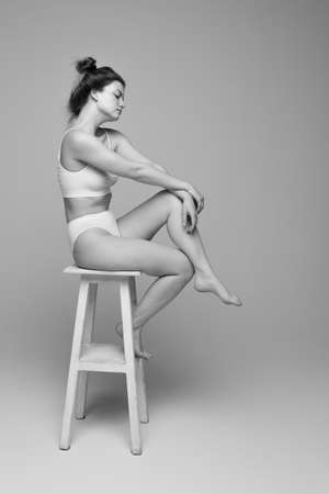 Studio portrait of young beautiful woman with slim body sitting on chair, posing in underwear. Black and white photography. Concept of natural beauty, body and skin care, femininity, self-acceptanceの写真素材