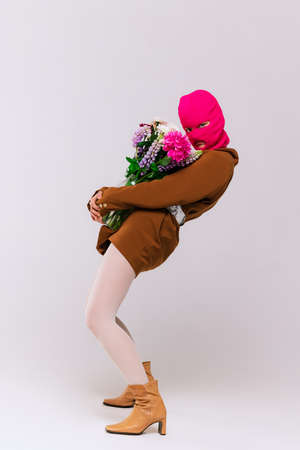 Portrait of stylish girl in brown jacket, white tights, boots and pink neon balaclava posing with flowers isolated over grey background. Concept of retro fashion, art photography, style, queer, beautyの写真素材