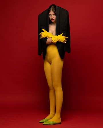 Portrait of young girl in oversized black jacket and yellow tights posing isolated over red background. Extraordinary fashion. Concept of retro fashion, art photography, style, queer, beautyの写真素材