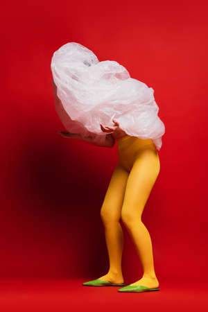 Portrait of young girl in yellow tights with cellophane covering face isolated over red background. Weirdness. Unusual art. Concept of retro fashion, art photography, style, queer, beautyの写真素材