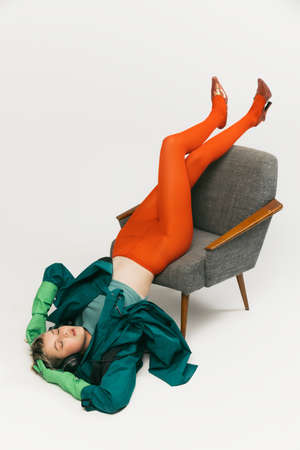 Portrait of stylish young girl in green coat and bright orange tight falling down the chair, posing isolated on grey studio background. Concept of retro fashion, art photography, style, queer, beautyの写真素材
