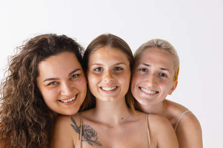 Portrait of three beautiful women posing together, smiling isolated over grey studio background....
