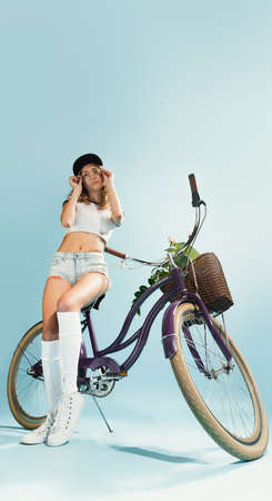 Portrait of beautiful young girl sitting on vintage bike, posing isolated over blue studio background. Spending good time. Concept of summer, fashion, retro style, emotions, freedom, funの写真素材