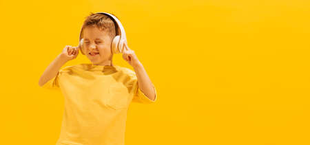 Portrait of cheerful little boy, child in bright T-shirt listening to music in headphones, posing isolated over yellow background. Concept of childhood, family, emotions, lifestyle, fashion, adの写真素材